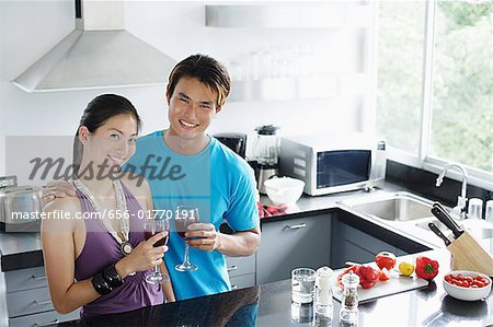 Couple standing in kitchen, holding wine glasses, looking at camera