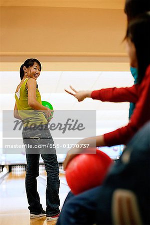 Three women at a bowling alley, one woman with bowling ball, looking over shoulder