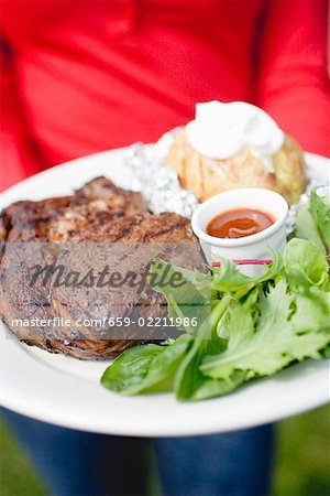 Person holding plate of grilled steak, baked potato & salad