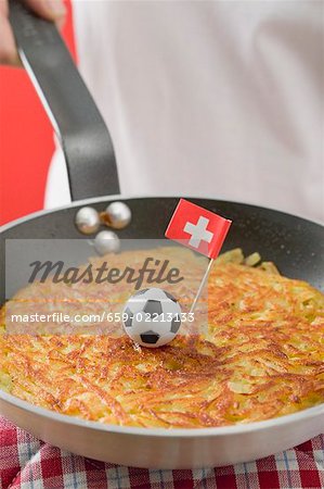 Hand holding frying pan with rösti, toy football & Swiss flag