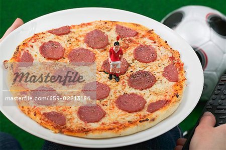 Hands holding salami pizza with toy footballer and remote