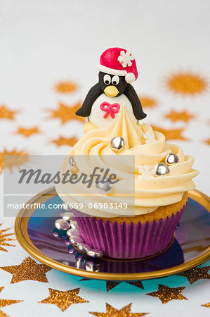 still life of a Christmas marzipan penguin sitting on top of a cream cup cake with gold star pattern on table