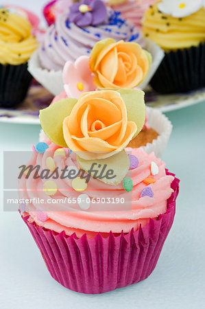 still life of a row of coloured cup cakes decorated with orange rose flowers on top in their cake papers on a white table with other cup cakes in the background