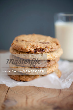 Chocolate chip, peanut butter and oatmeal cookies stacked on baking parchment wih a glass of milk in the background.