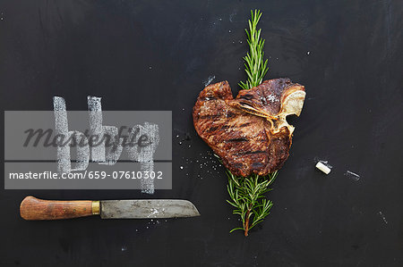 A grilled T-bone steak with rosemary and knife on a chalkboard