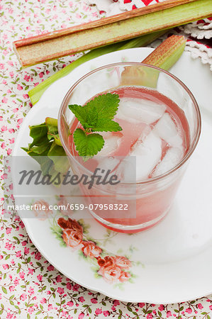 A glass of rhubarb spritzer with ice cubes and pieces of rhubarb on an old, rose patterned plate
