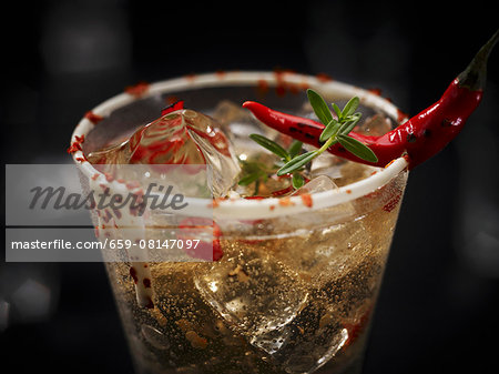 A champagne drink with chilli and ice cubes garnished with a chilli pepper and herbs in glass with a white chocolate rim