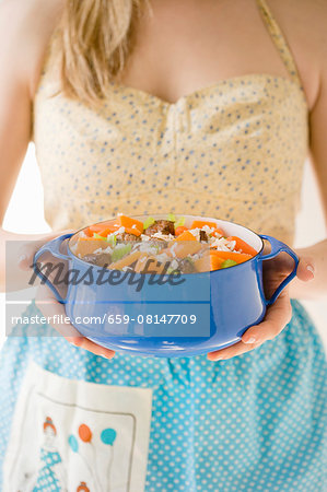 A woman wearing a dress and an apron serving a beef stew with carrots, celery and rice