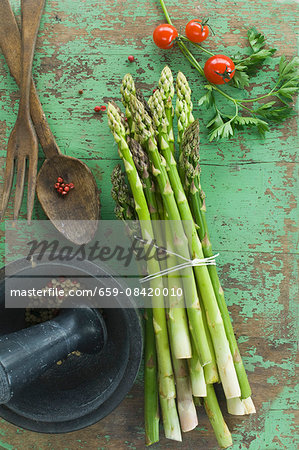 A bundle of fresh green asparagus, tomatoes and parsley on a wooden table, peppercorns in a mortar and salad servers A bundle of fresh green asparagus, tomatoes and parsley on a wooden table, peppercorns in a mortar and salad servers