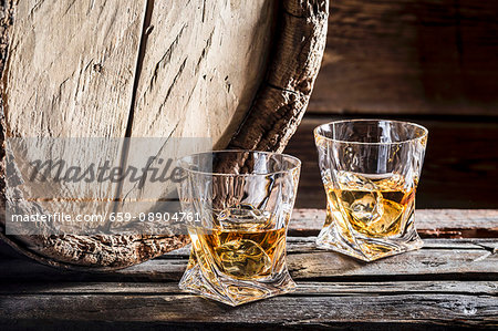 Two glasses of whiskey on the rocks in front of an old wooden barrel