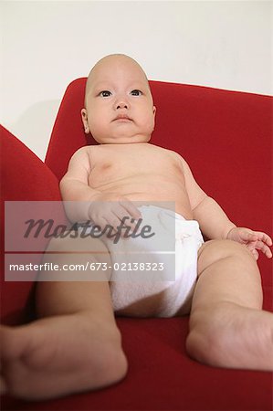 A baby seated on a red sofa and wearing a diaper.