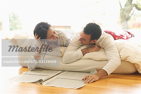 A couple lounge in bed while reading the newspaper.