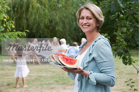 Woman with watermelon slices in garden, family members in background