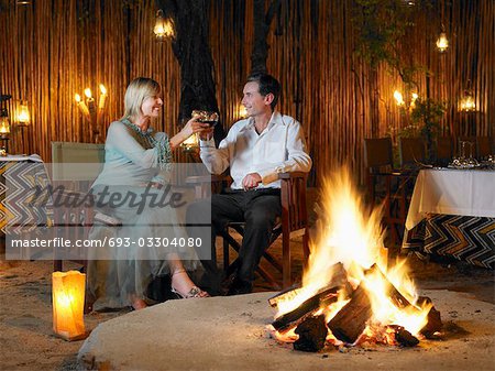 Couple sitting on chairs at outdoor nightclub near bonfire, toasting
