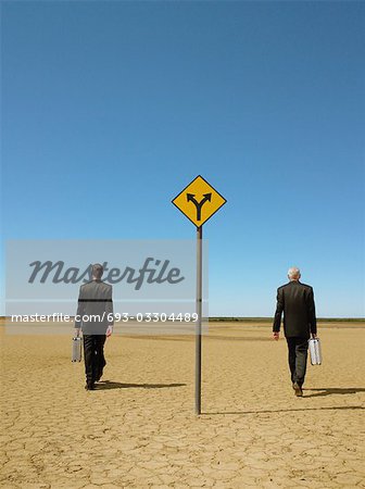 Two businessmen with briefcases walking past road sign in desert, back view