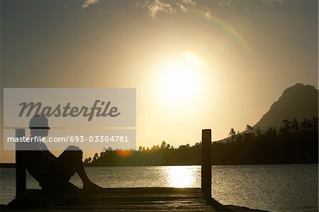 Man sitting on dock by lake, side view.