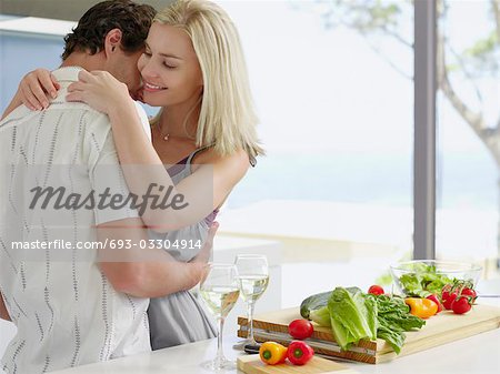 Young couple hugging in kitchen near countertop with fresh produce and wineglasses