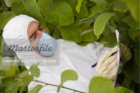Worker in protective mask and suit writing on pad amongst plants