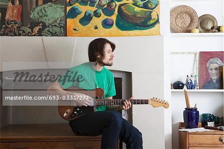 Young man sits in living room playing electric guitar