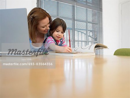 Girl (3-4) colouring in book beside mother using laptop, in home