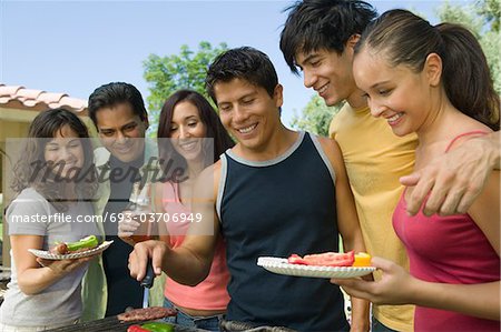 Group of young people gathered around grill at outdoor picnic.
