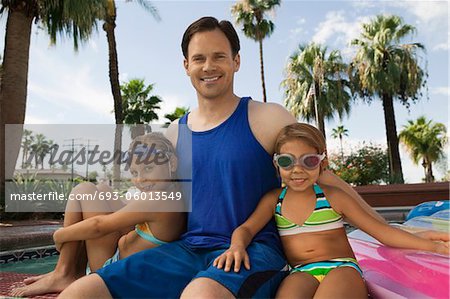 Two girls (7-9) sitting with father by pool, portrait.