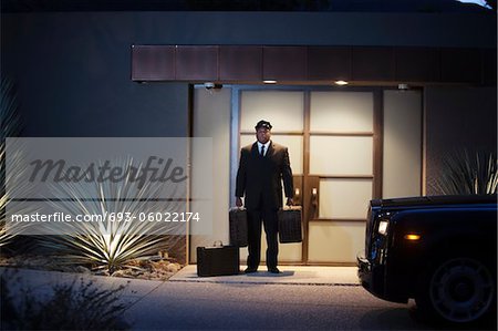 Chauffeur stands at lit entrance doorway with luggage