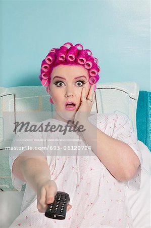 Portrait of shocked woman holding remote with hair curlers sitting on bed