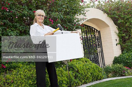 Portrait of a happy senior female agent hammering sign board in lawn