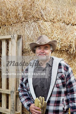 Portrait of a happy mature man with cowboy hat holding gloves in front of hay stack