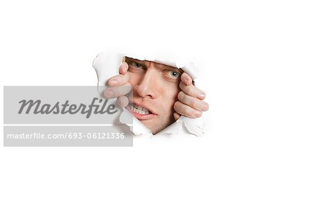 Portrait of a aggressive mid adult man peeking from ripped white paper hole