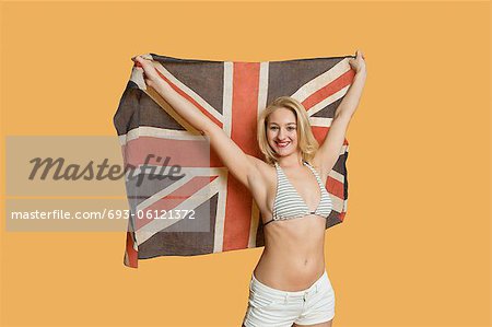 Portrait of a beautiful young woman holding British flag with arms raised over colored background