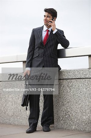 Young Indian businessman using cell phone with laptop bag by parapet