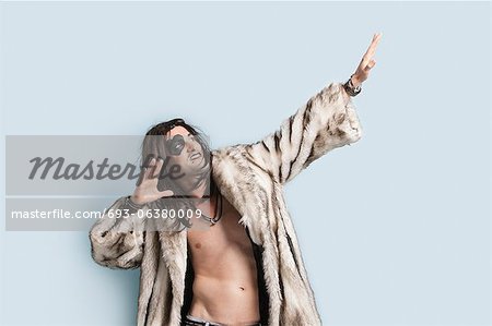 Young man in fur coat looking up with arms raised against light blue background