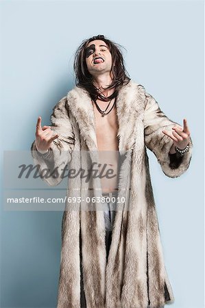 Portrait of young man in fur coat gesturing rock music sign against light blue background