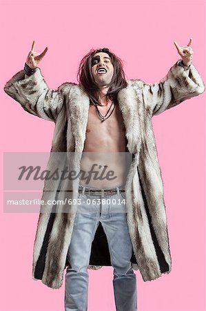 Young man in fur coat gesturing rock music sign over pink background