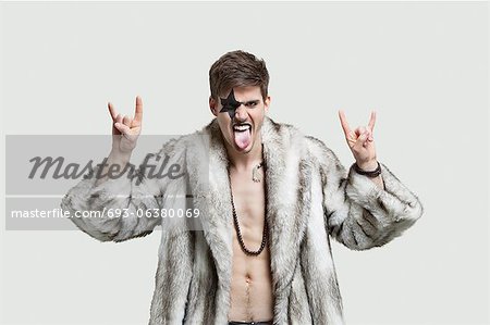 Portrait of an angry young man making rebellious gestures and sticking out tongue against gray background