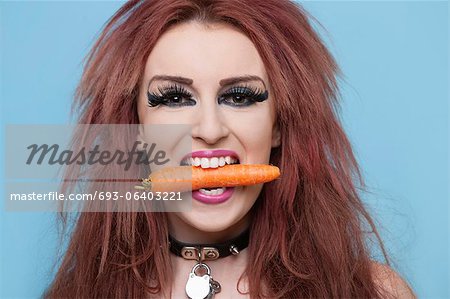 Portrait of funky young woman holding carrot in mouth over blue background