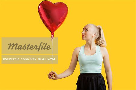 Beautiful young woman looking at heart shaped balloon over yellow background