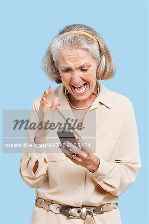 Irritated senior woman reading text message on cell phone against blue background