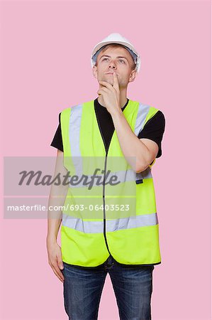 Thoughtful young construction worker looking away over pink background