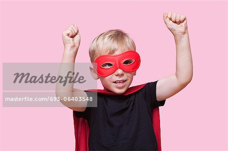 Young boy dressed in superhero costume with arms raised over pink background