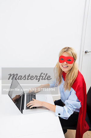 Portrait of happy businesswoman in superhero costume working on laptop in office