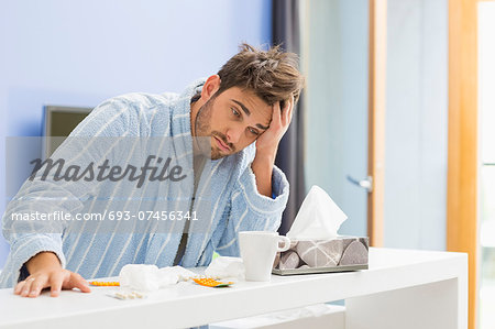 Young ill man with coffee mug, medicine and tissue leaning on kitchen counter