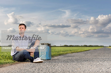 Full length of young man with empty gas can sitting by country road