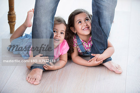 Playful girls holding father's legs on hardwood floor