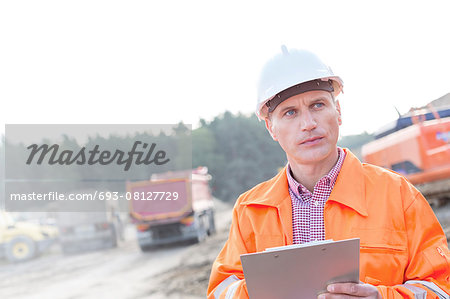 Engineer looking away while holding clipboard against clear sky