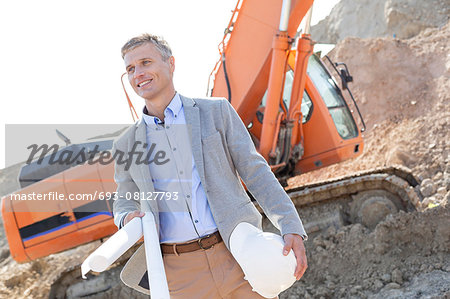 Smiling architect looking away while holding blueprints and hardhat at construction site