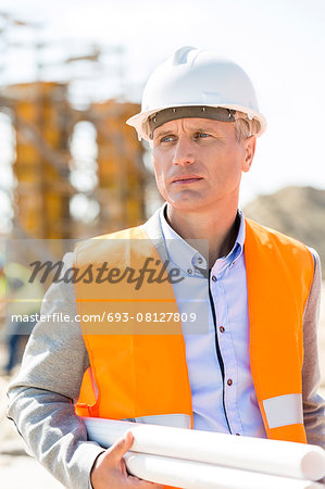 Thoughtful architect looking away while holding blueprints at construction site