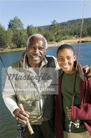 Grandfather and grandson holding fishing rods by lake, smiling, (portrait)
